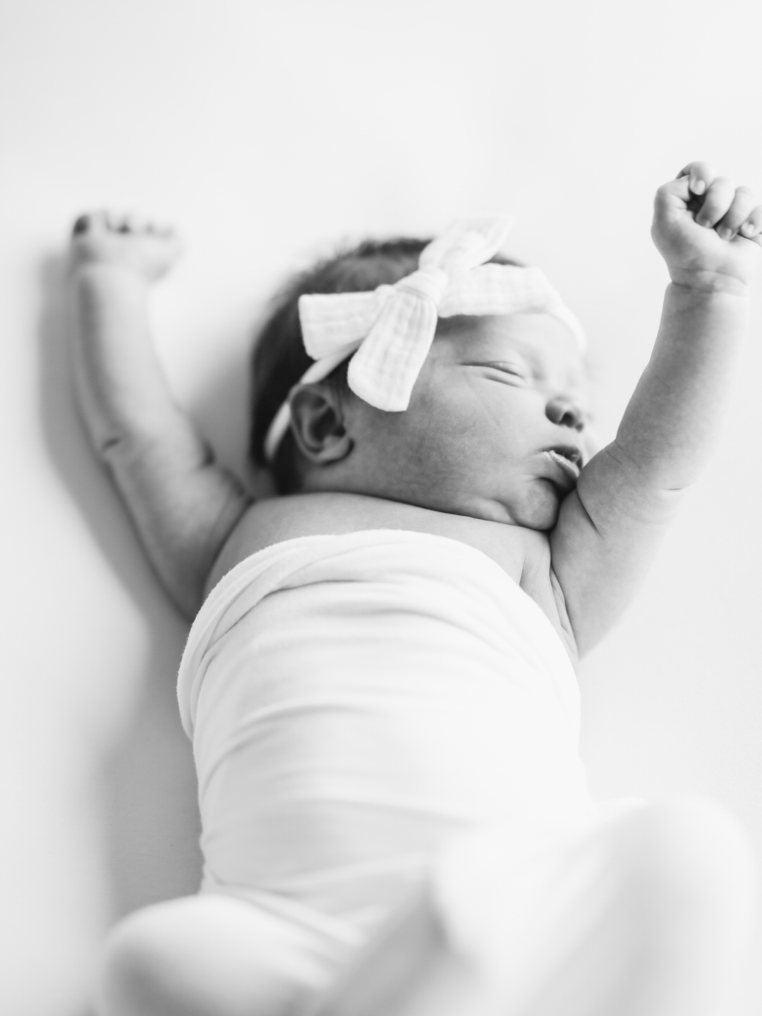 newborn baby stretching during photo session in studio. wearing white bow and wrapped in soft white swaddle.