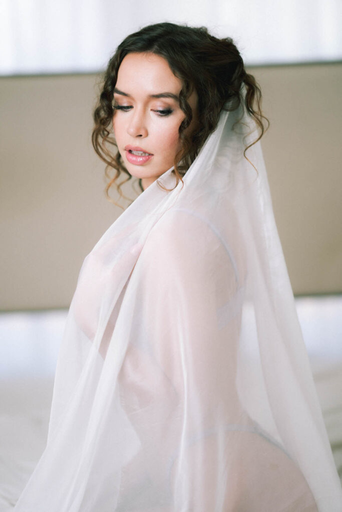 bride in her veil taking boudoir images on a white bed for her soon to be husband. professional portrait transforming self image