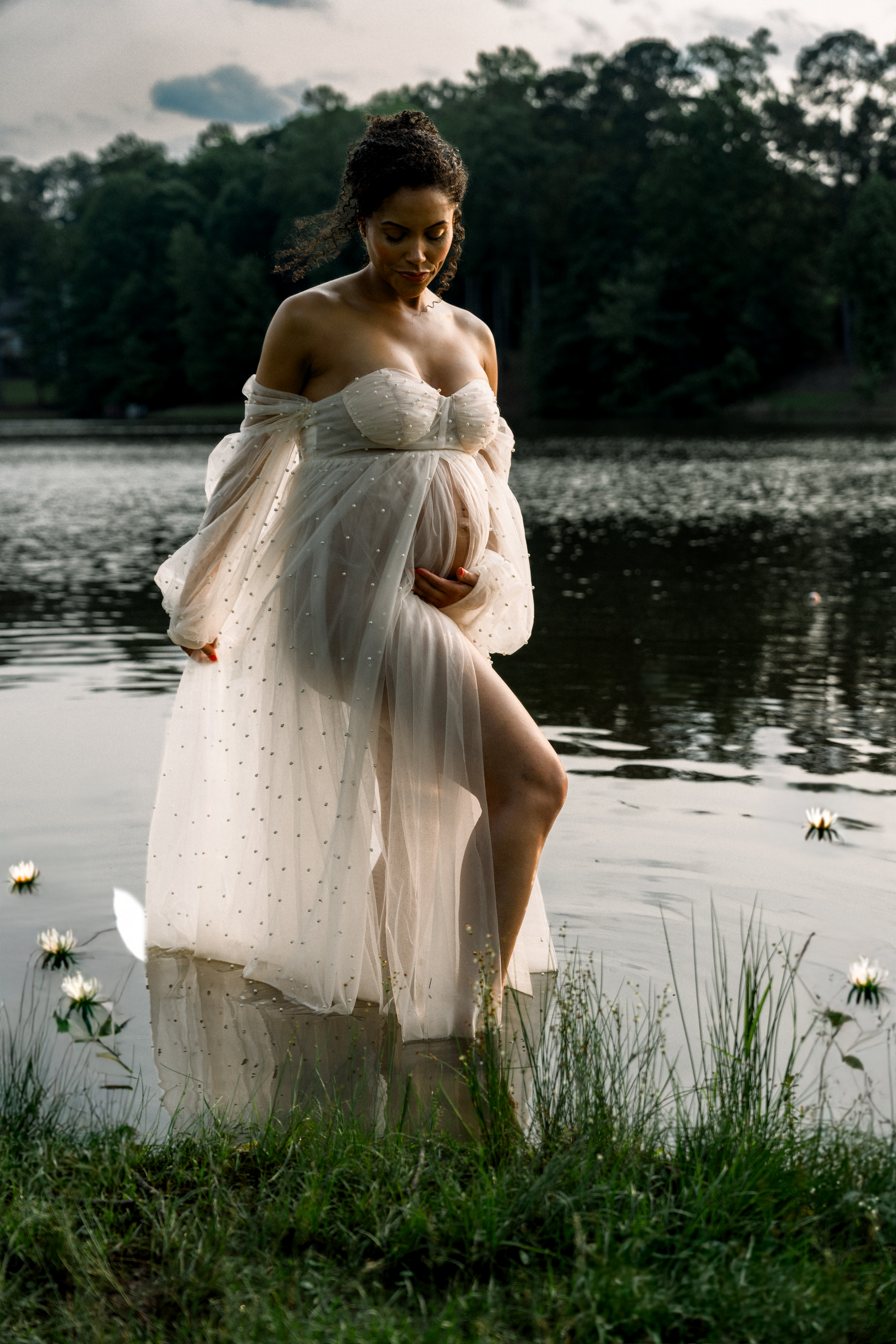 woman standing in a calm lake in a sheer lace and pearl maternity down showing her pregnant belly