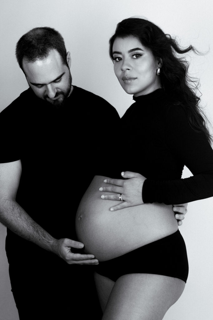 woman posing with her pregnant belly and husband is rubbing her belly in black and white photograph