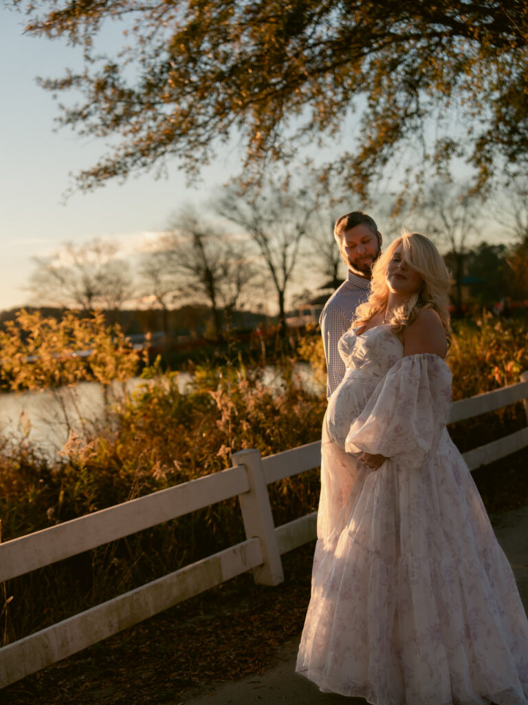 Woman standing with husband in flowing maternity gown showing her bump with sunset in background of lake