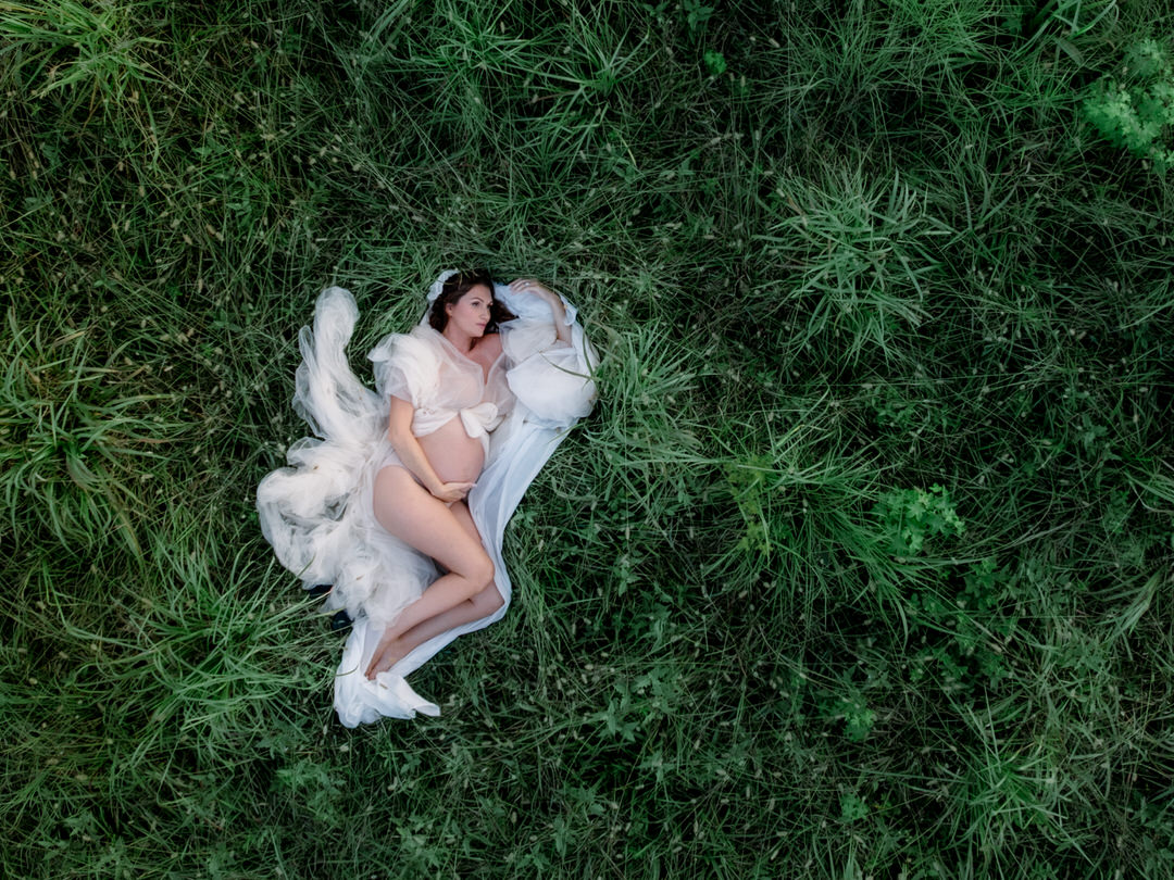 aerial photograph of woman laying in a green field in a maternity gown holding her pregnant belly