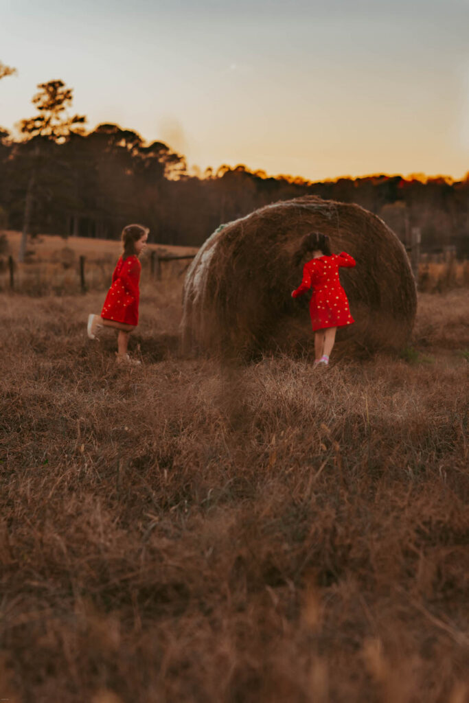 two young girls playing near a hay bail on a farm field during sunset