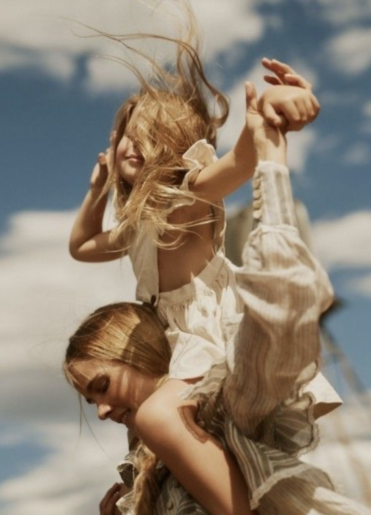 young girl sitting on moms shoulders wind blowing their hair bright blue sky