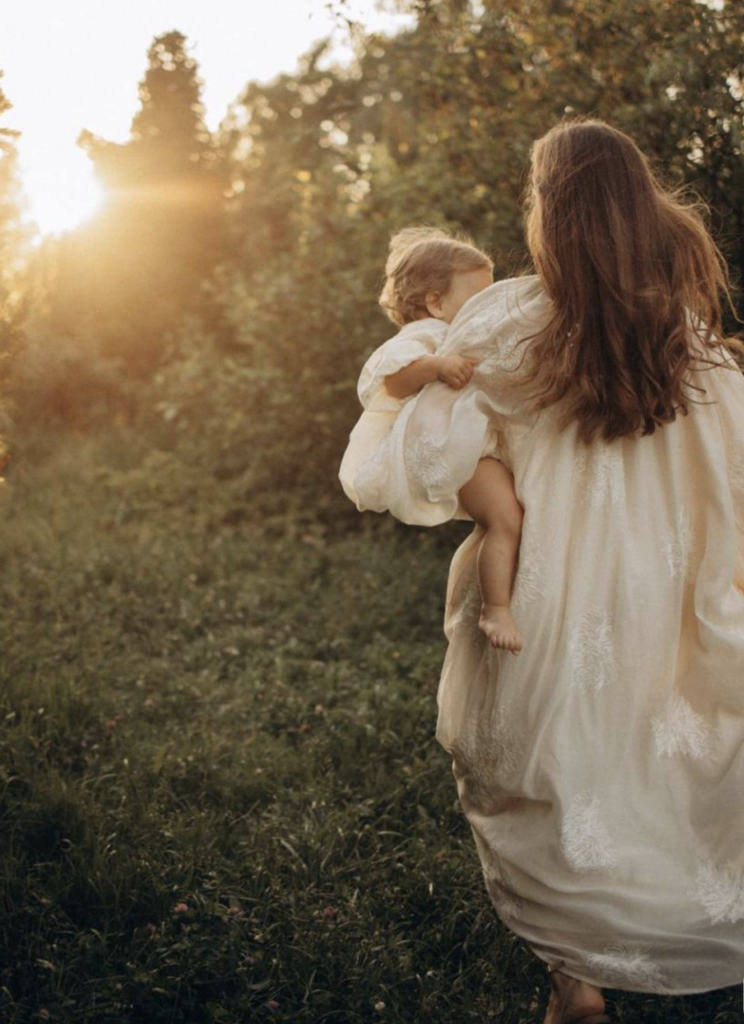 mom walking towards sunset in big flowing white gown holding her baby