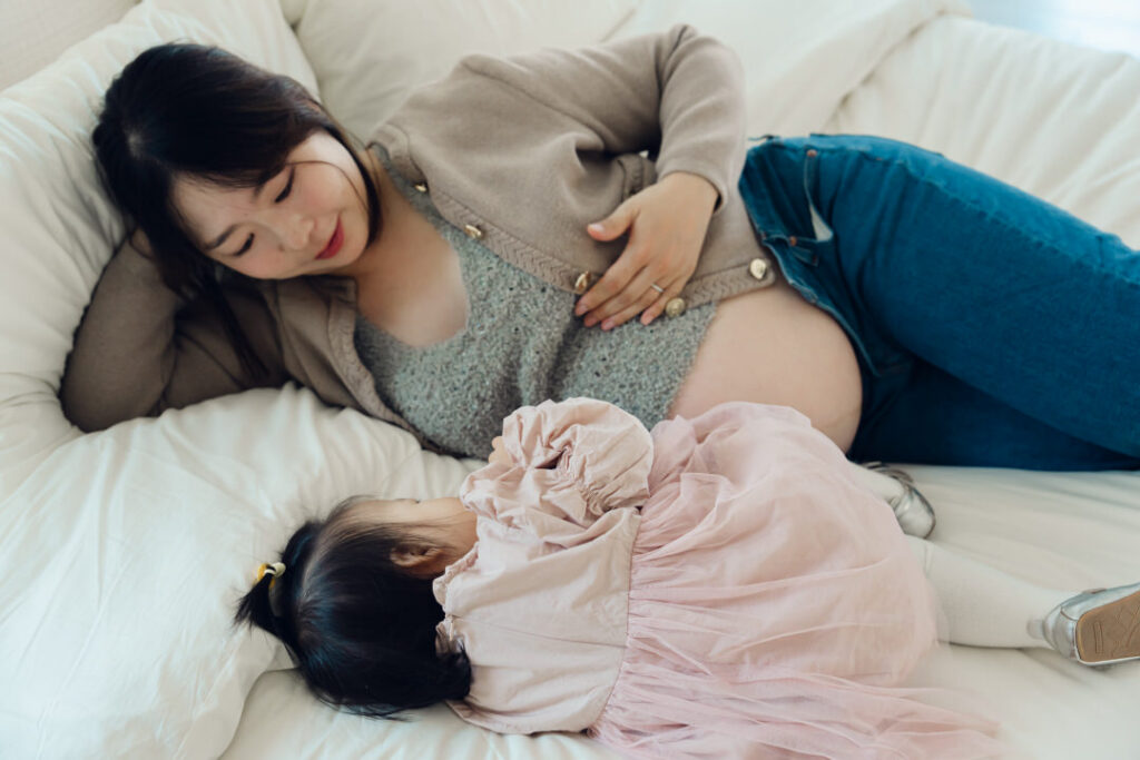 pregnant korean mom touching belly while laying bed with small toddler girl